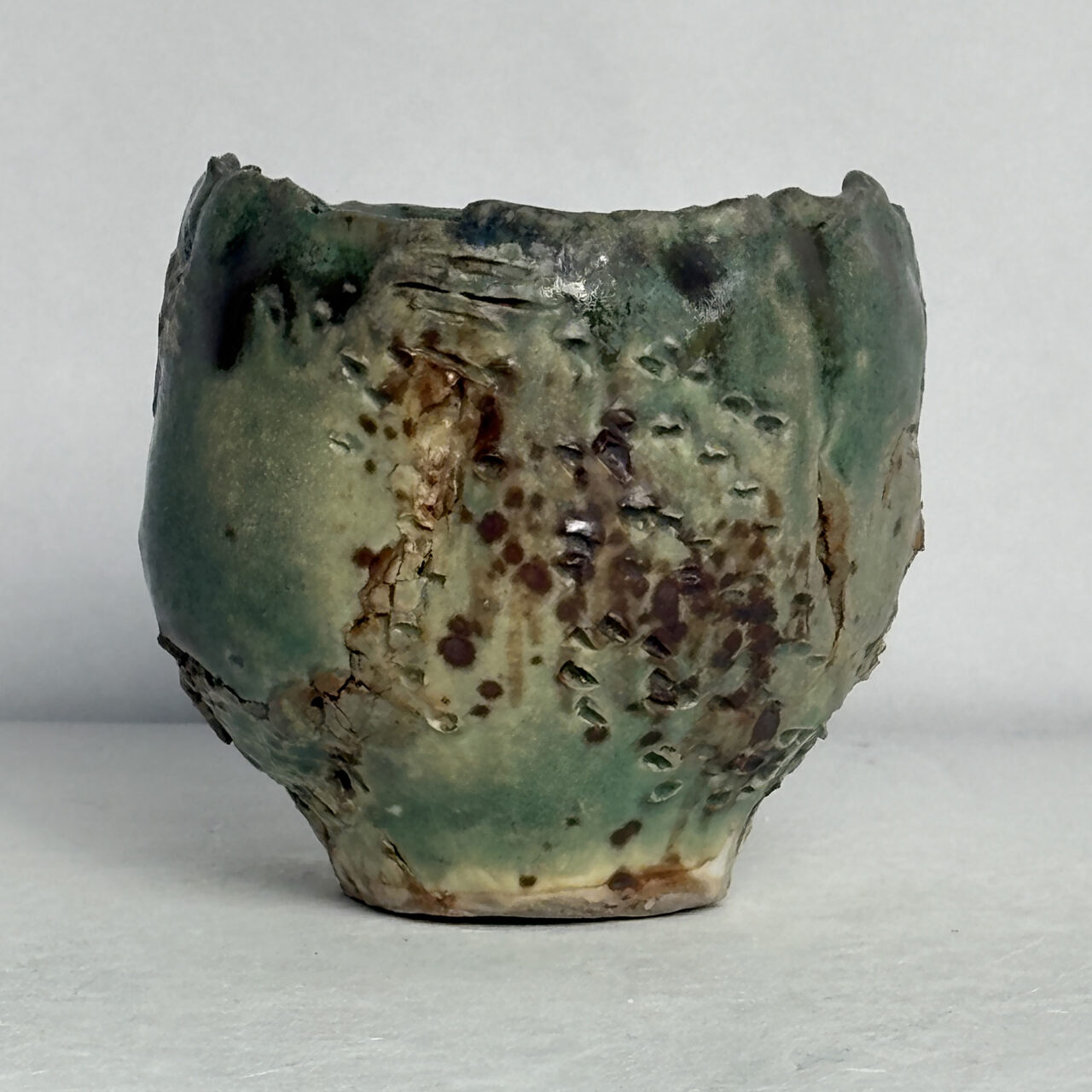 No. 100 Small ceramic vessel with matte green glaze, textured surface, and scattered rust and dark brown markings.