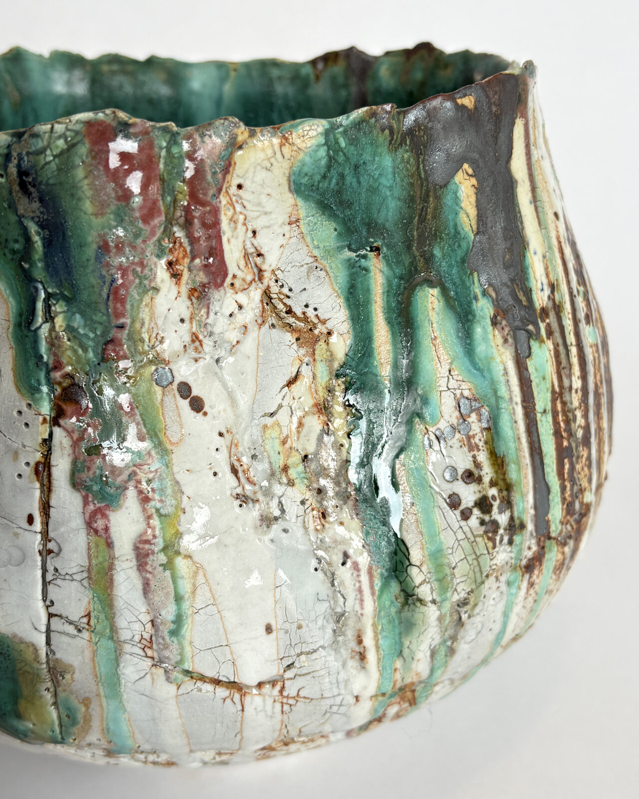 Close-up of a sculptural ceramic vessel with flowing emerald green, turquoise, and deep red glazes over cracked white porcelain slip and dark iron markings.