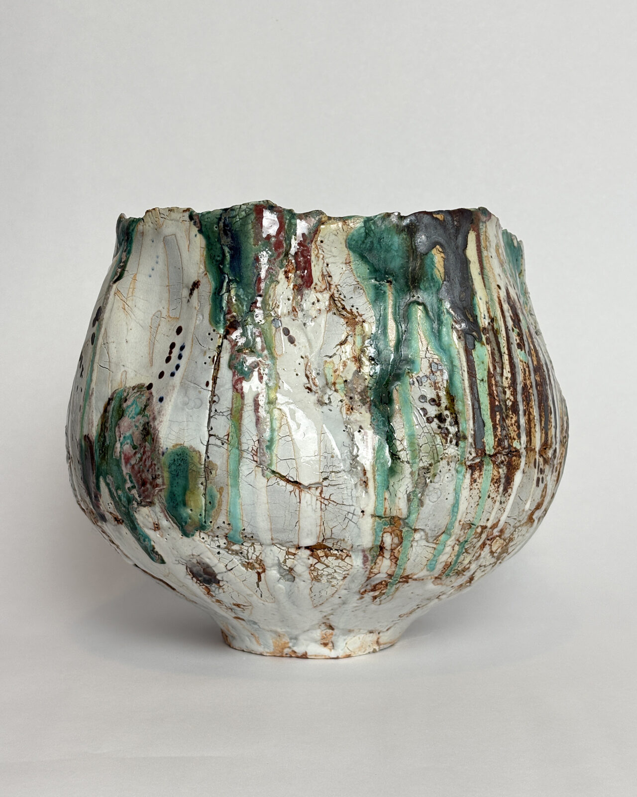 Front view of a hand-built ceramic vessel with a rounded body, narrow base, irregular rim, and vertical green and red glaze drips over textured white clay.