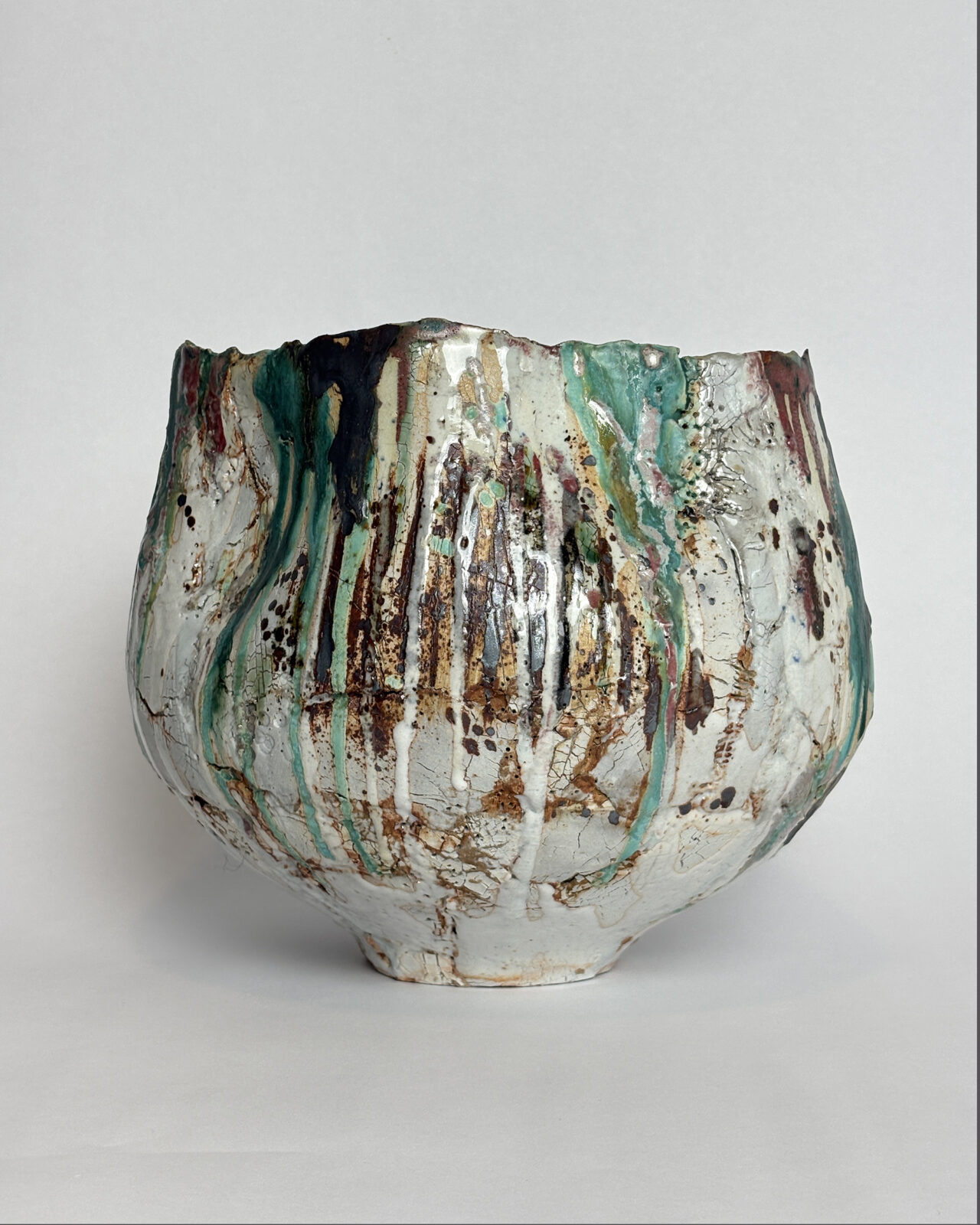 Side view of a sculptural ceramic vessel featuring long streaks of turquoise, dark green, and rust-colored glazes over a fractured white surface.