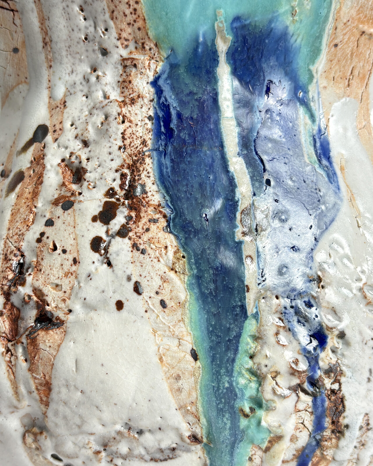 Extreme close-up of a ceramic surface showing layered turquoise and cobalt blue glazes flowing over cracked white porcelain slip, rust-colored iron deposits, and pitted texture.