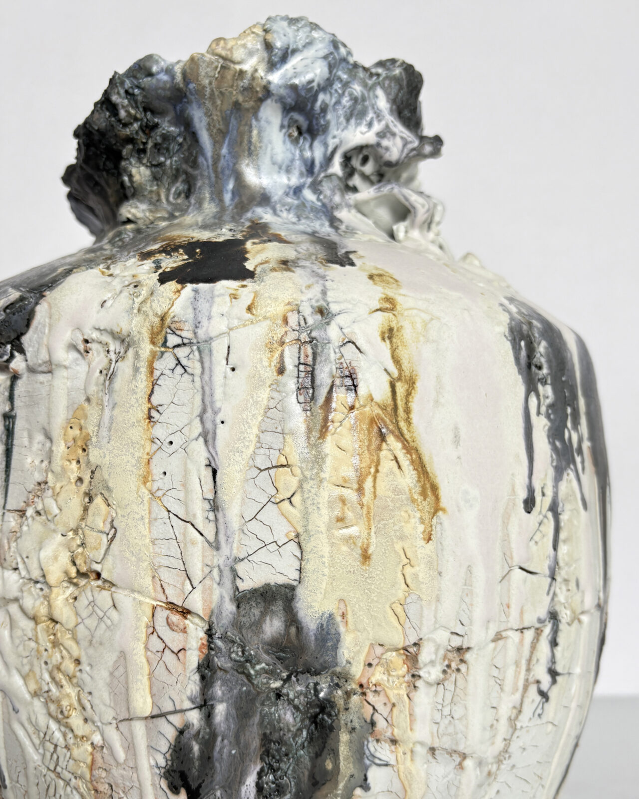 Close-up of a sculptural ceramic vessel with cracked white porcelain slip, ochre markings, flowing charcoal-gray glazes, and a rugged, heavily textured rim.
