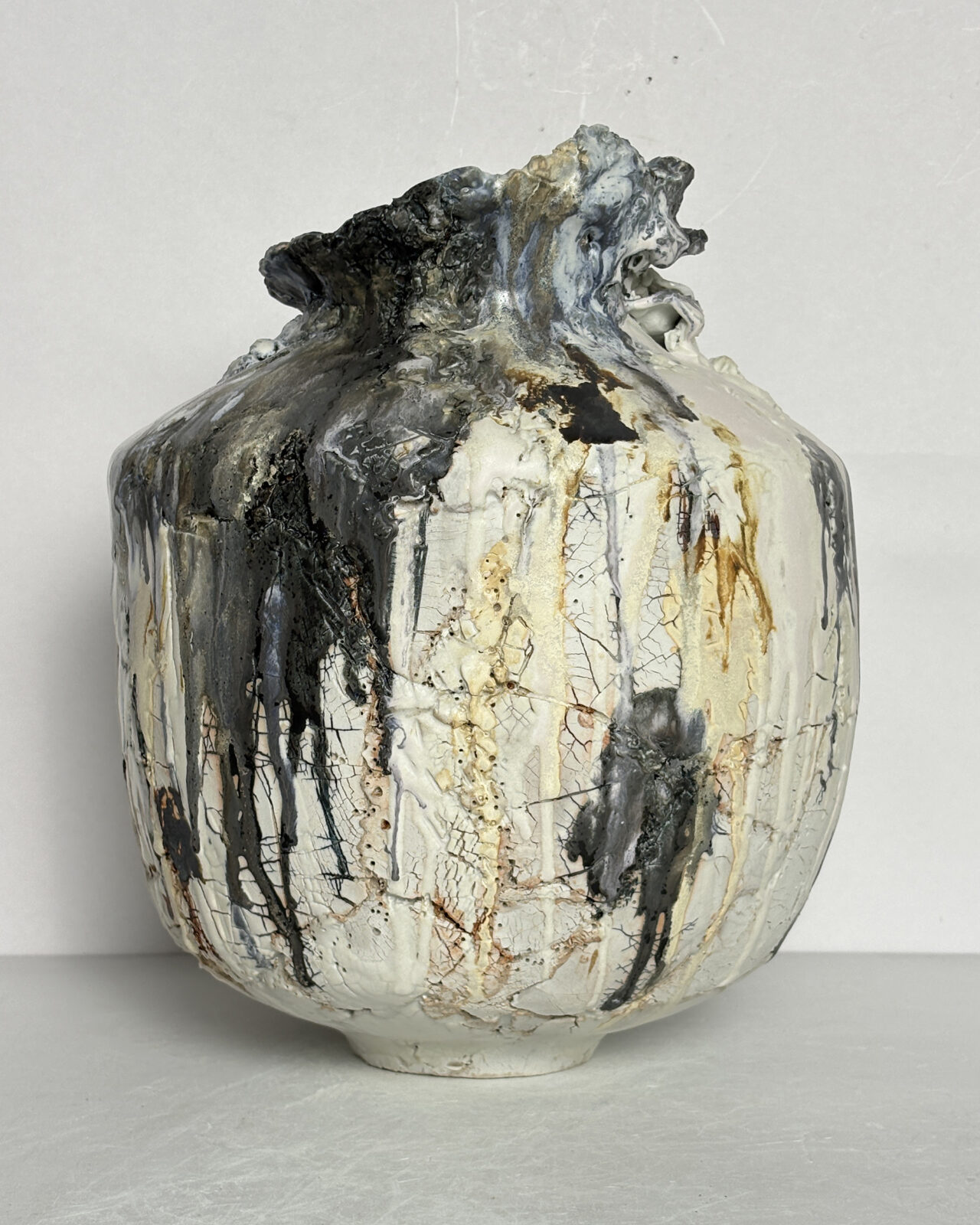 Front view of a hand-built ceramic vessel with broad angular shoulders, a narrow neck, and a dramatic sculptural rim. Fractured white surfaces contrast with dark gray glaze drips and warm ochre tones.