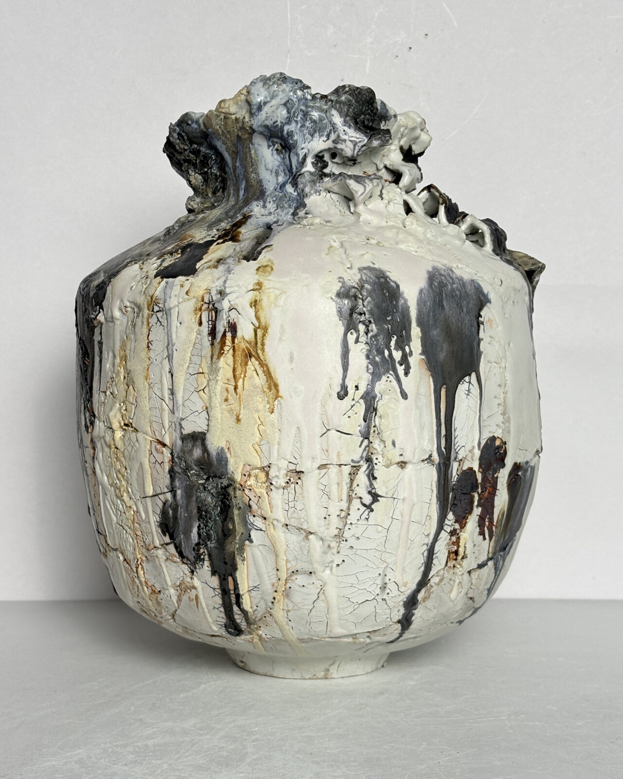 Side view of a ceramic vessel with a wide, structured body, narrow neck, rugged rim, and bold charcoal-gray glaze drips over cracked white porcelain slip.