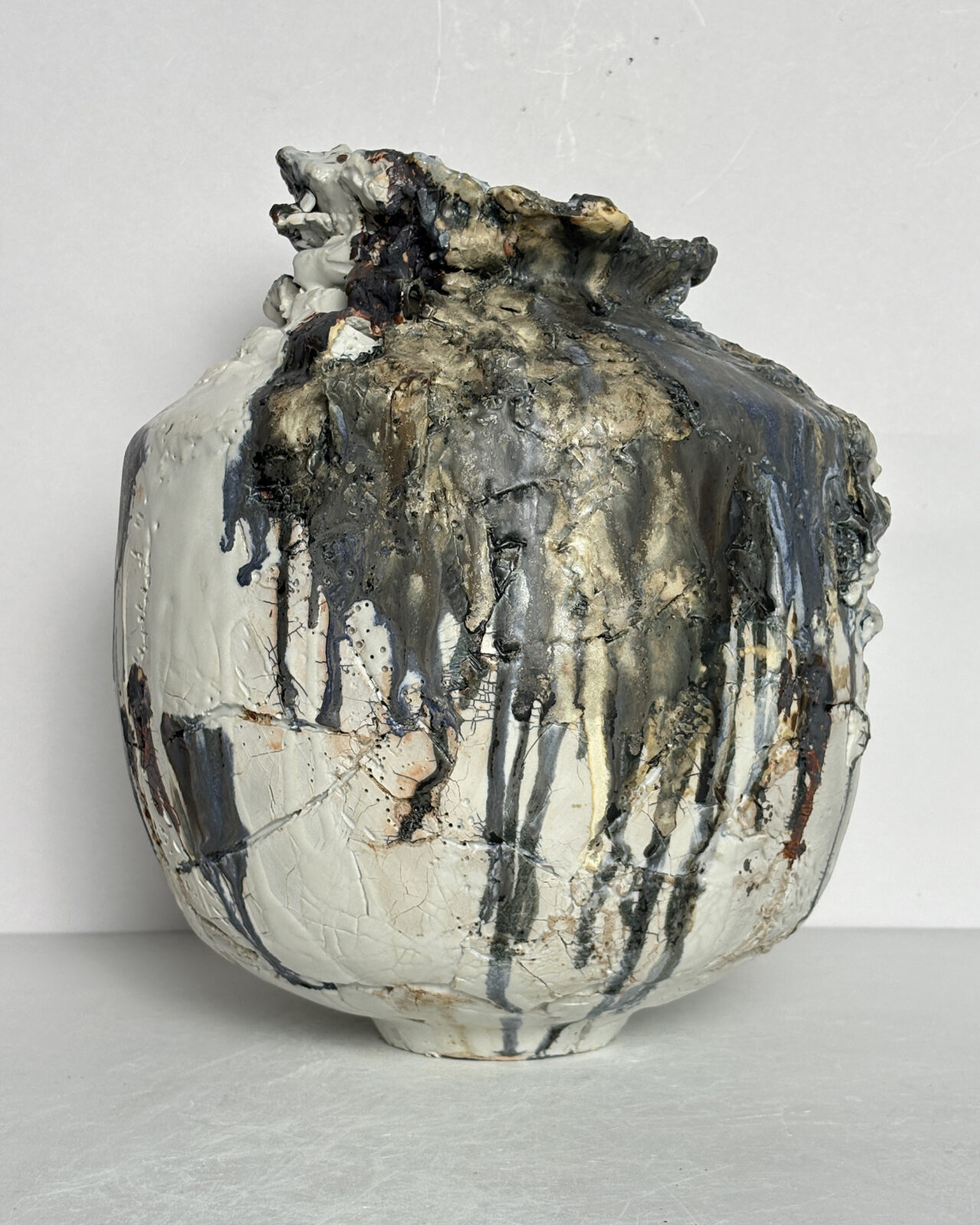Sculptural ceramic vessel featuring pronounced shoulders, a narrow opening, and heavy gray glaze buildup flowing down one side over a pale, fractured surface.