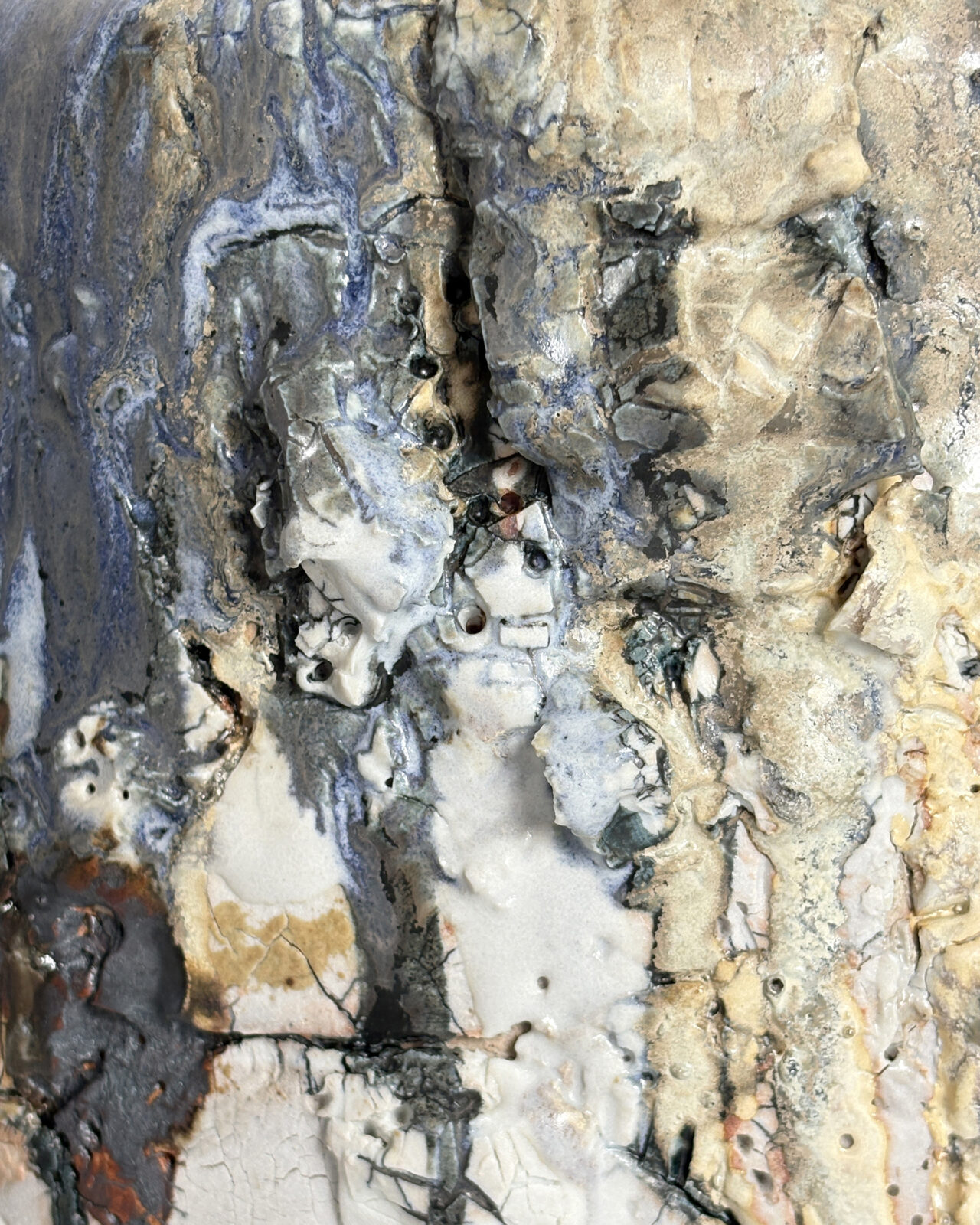 Extreme close-up of a ceramic surface showing rough clay formations, fractured porcelain slip, layered mineral-gray and blue glazes, and areas of glossy and matte texture.