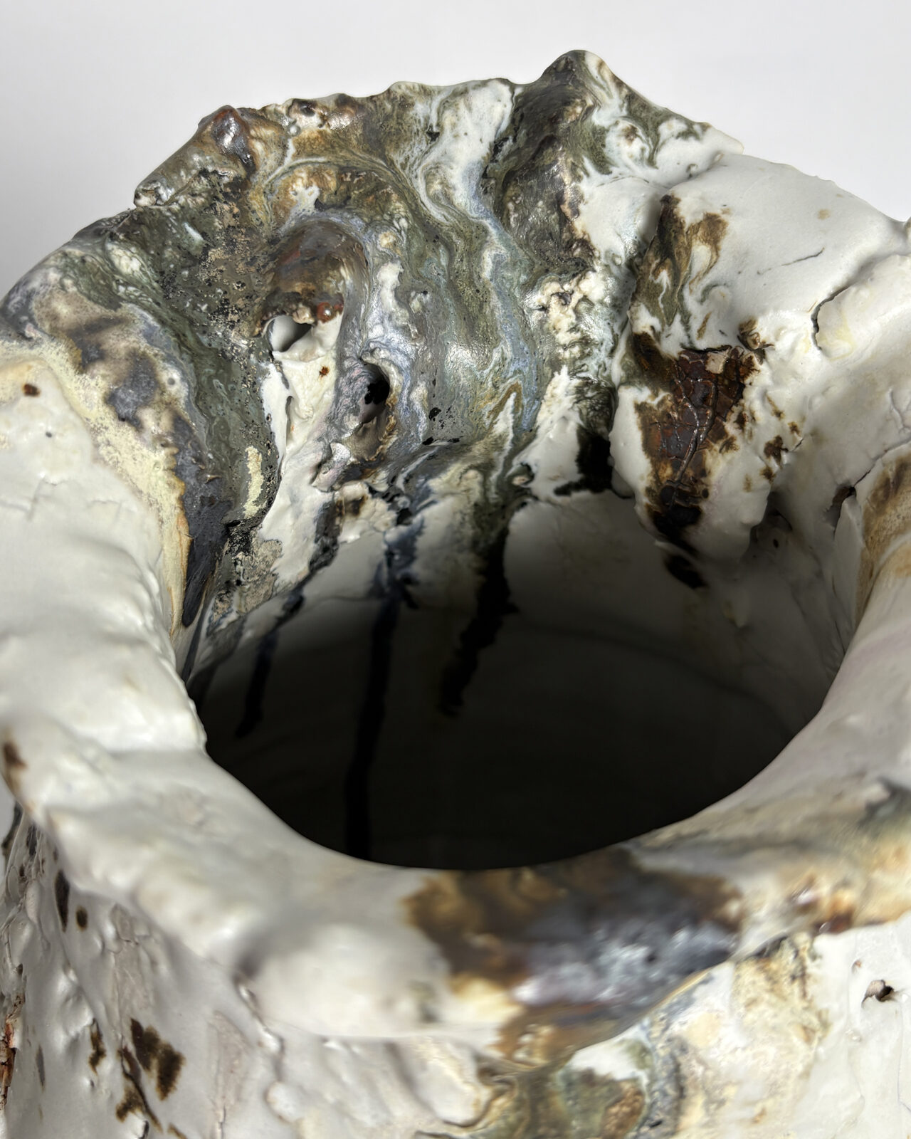 Overhead close-up of an asymmetrical ceramic vessel rim with folded clay formations, small openings, and swirling metallic gray, cream, and earthy green glazes.