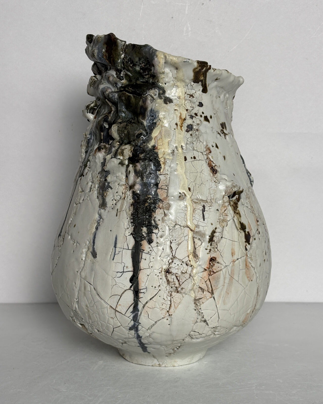 Front view of a hand-built ceramic vessel with a rounded body, narrow neck, and dramatic sculptural rim, featuring cracked white surfaces and dark gray glaze drips.