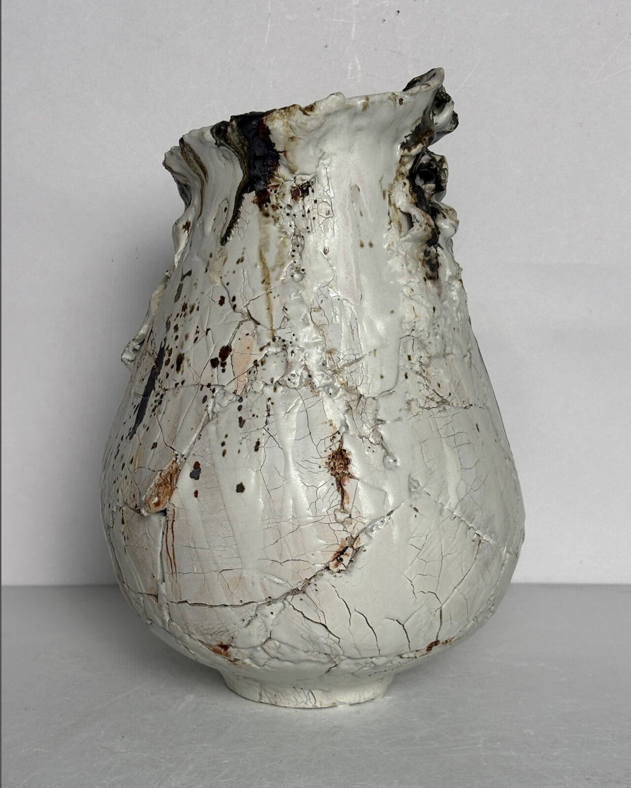 Side view of a ceramic vessel with a wide, rounded base, gently tapered neck, and irregular rim, showing pale fractured porcelain slip and scattered iron oxide markings.