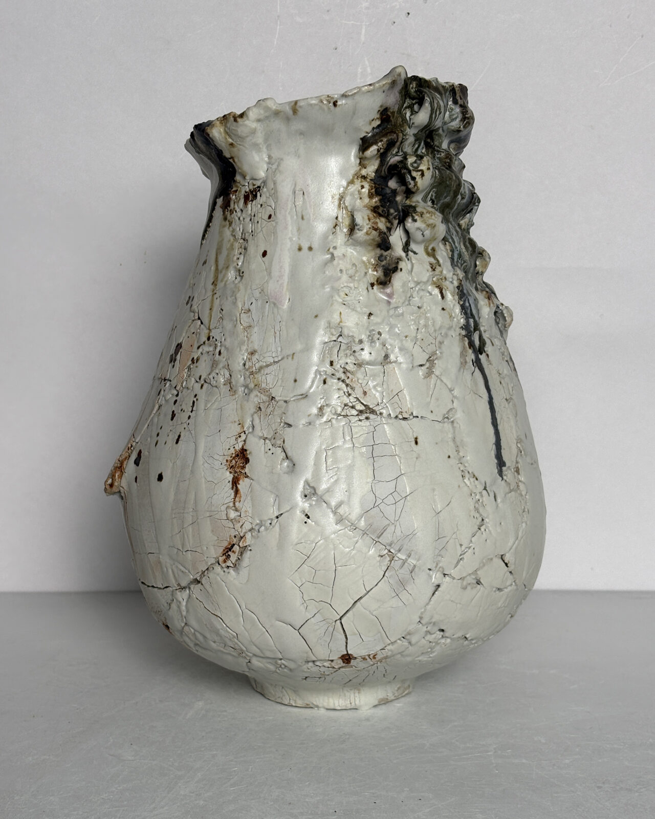 Sculptural ceramic vessel with a pale rounded body, asymmetrical rim, and heavily built-up clay formations along one side, accented by metallic gray glazes and subtle ochre tones.