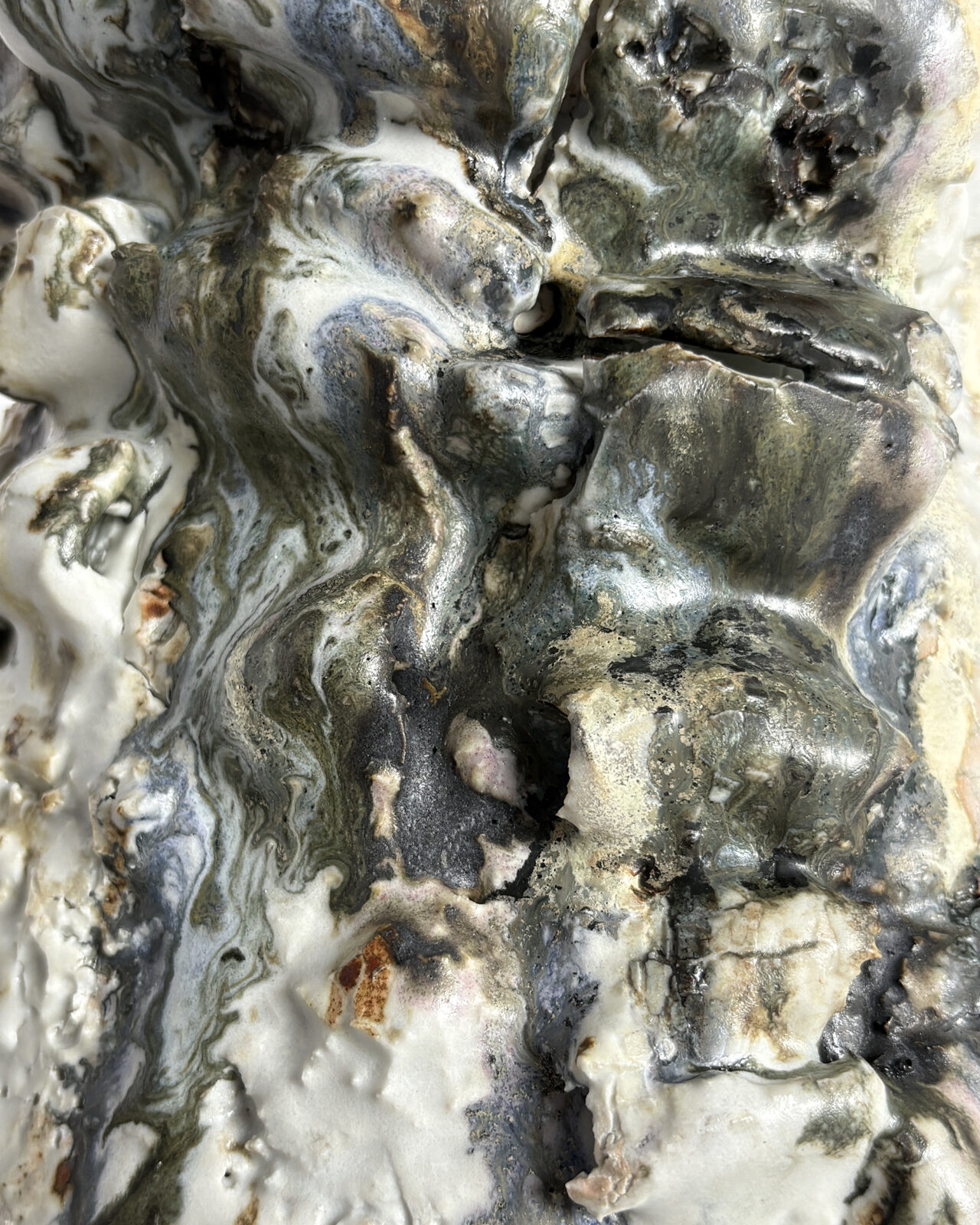 Extreme close-up of layered ceramic textures showing folded and built-up clay, glossy metallic gray and cream glazes, small cavities, and rough geological surfaces.